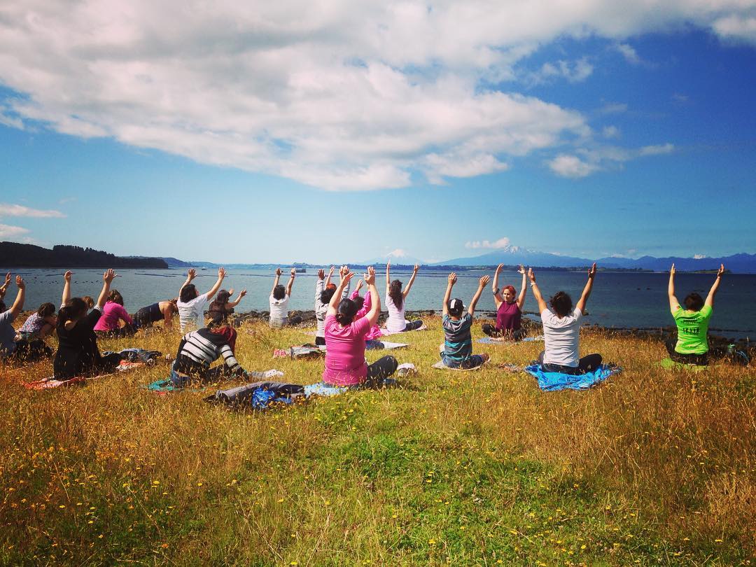 Un grupo de personas realizando yoga en un espacio al aire  libre, al fondo la vista al lago llanquihue y los volcanes.